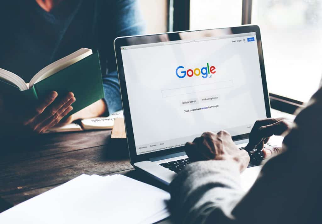 Página do Google exibida em um notebook, que está em cima de uma mesa; há uma pessoa teclando nele e, à sua frente, há uma pessoa lendo um livro
