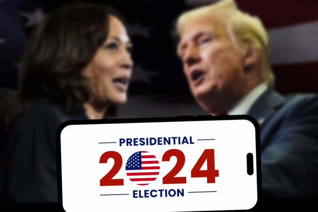 Rostos de Harris e Trump desfocados, com a frase 2024 Presidential Election sendo exibida à frente em um smartphone