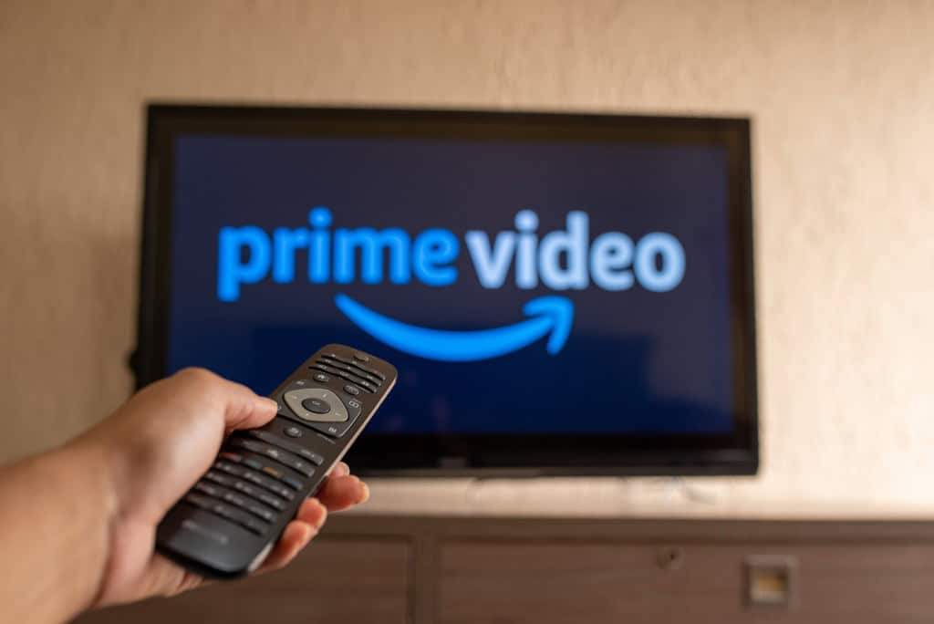Logo do Prime Video em uma TV ao fundo, desfocada; à frente, uma pessoa com um controle remoto de TV na mão