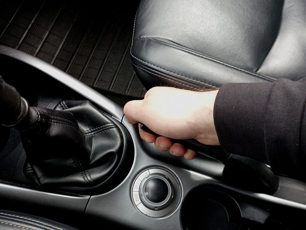 Um homem engatando o freio de mão de um carro. Dirigir com o pedal do freio travado ou com o freio de mão engatado são fatores que danificam os pedais do carro
