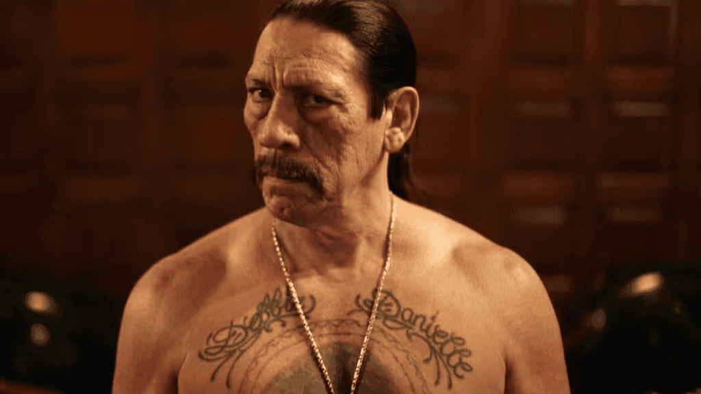 Danny Trejo com expressão séria aparece sem camisa, exibindo tatuagens no peito e usando um colar comprido, em cena de estilo intenso ligada ao tema “atores que mais morreram em filmes e séries”.