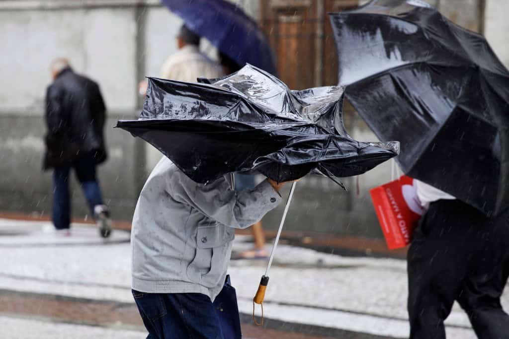 Pessoa segura um guarda-chuva virado pela força do vento durante um temporal, enquanto outras caminham sob chuva ao fundo.